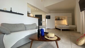 Una sala de estar moderna con un sofá gris, una mesa de centro de madera con una botella y un sombrero, y una cama al fondo.