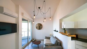 Una sala de estar moderna con un sofá, una mesa de centro y un televisor. Se observa una cocina con armarios y una encimera.