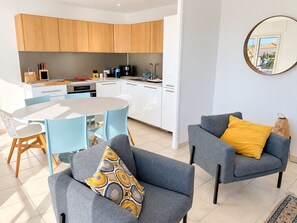A modern kitchen with white cabinets, a dining table with blue chairs, and a grey sofa with a yellow pillow.