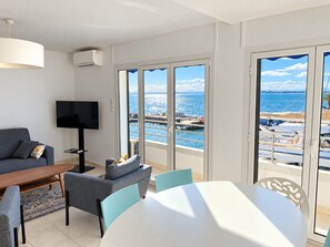 A modern living room with a flat-screen TV, a dining area with a table and chairs, and large windows offering a view of the sea and boats.