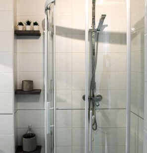A glass-enclosed shower with a handheld showerhead and a wall-mounted faucet. There are three shelves with decorative items and a small trash can.