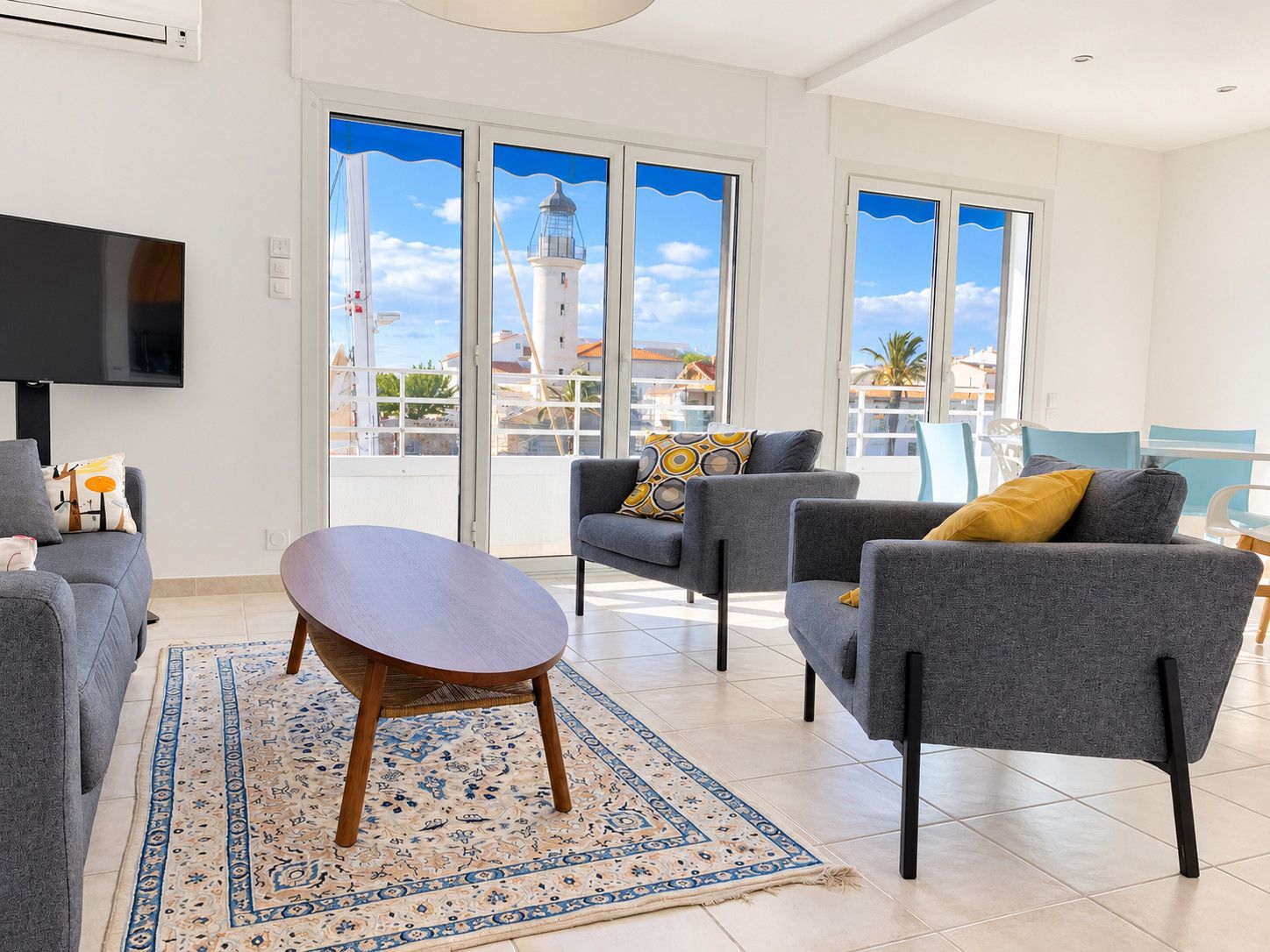 A modern living room with a flat-screen TV, grey sofas, a patterned rug, and large windows with a view of a lighthouse.
