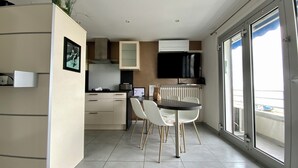 Une cuisine moderne avec des meubles blancs, une table à manger avec des chaises, un téléviseur fixé au-dessus du comptoir et une grande fenêtre donnant sur l’extérieur.