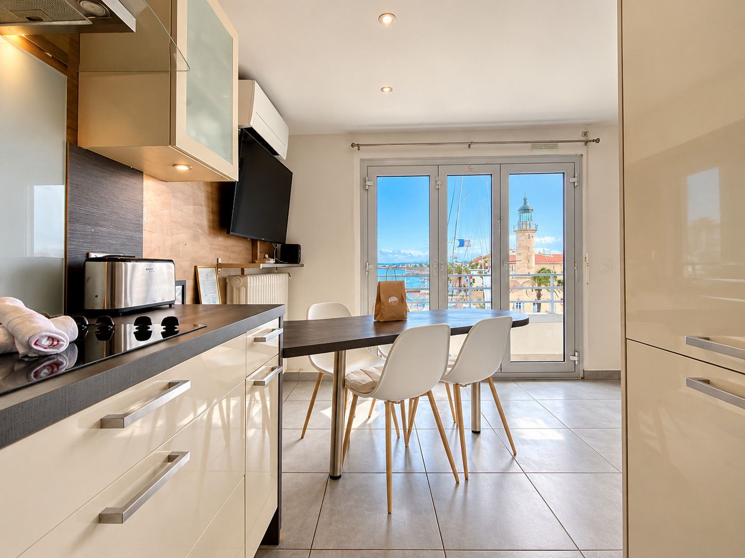 A modern kitchen with a dining area, a flat-screen TV, and a view of a coastal town.