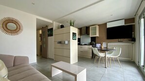 Un salon moderne avec un canapé, un coin repas avec des chaises, une cuisine équipée d’armoires et un téléviseur fixé au mur.