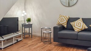 A modern living room with a grey sofa, two patterned cushions, a small side table with a potted plant, a round mirror on the wall, and a television on a stand.