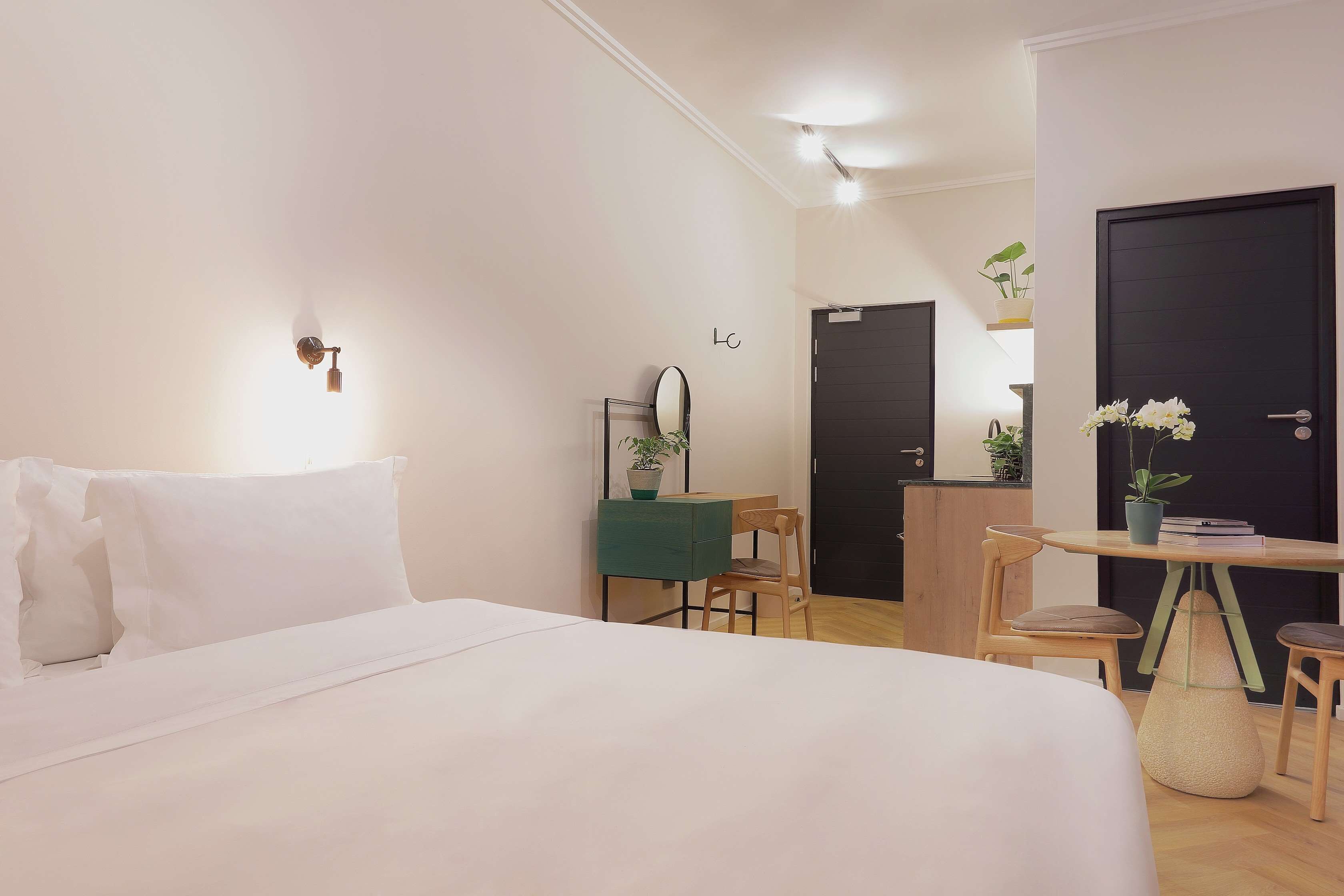 A modern hotel room with a bed, a desk with a chair, a small table with a potted plant, and a door.
