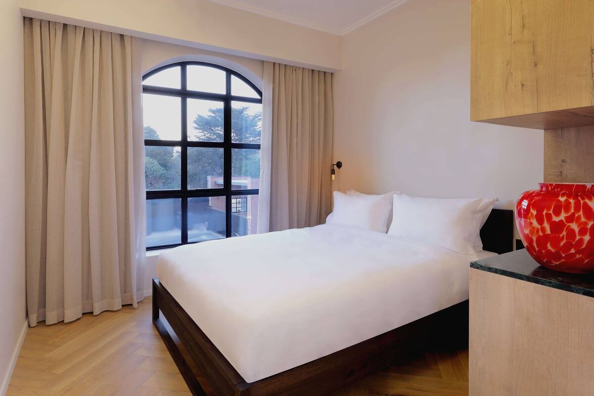 A hotel room with a large bed, a wooden headboard, a window with curtains, and a red vase on the nightstand.