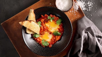Une assiette de shakshuka accompagnée de tranches de pain grillé et d’un petit bol de sel, le tout présenté sur un plateau en bois.