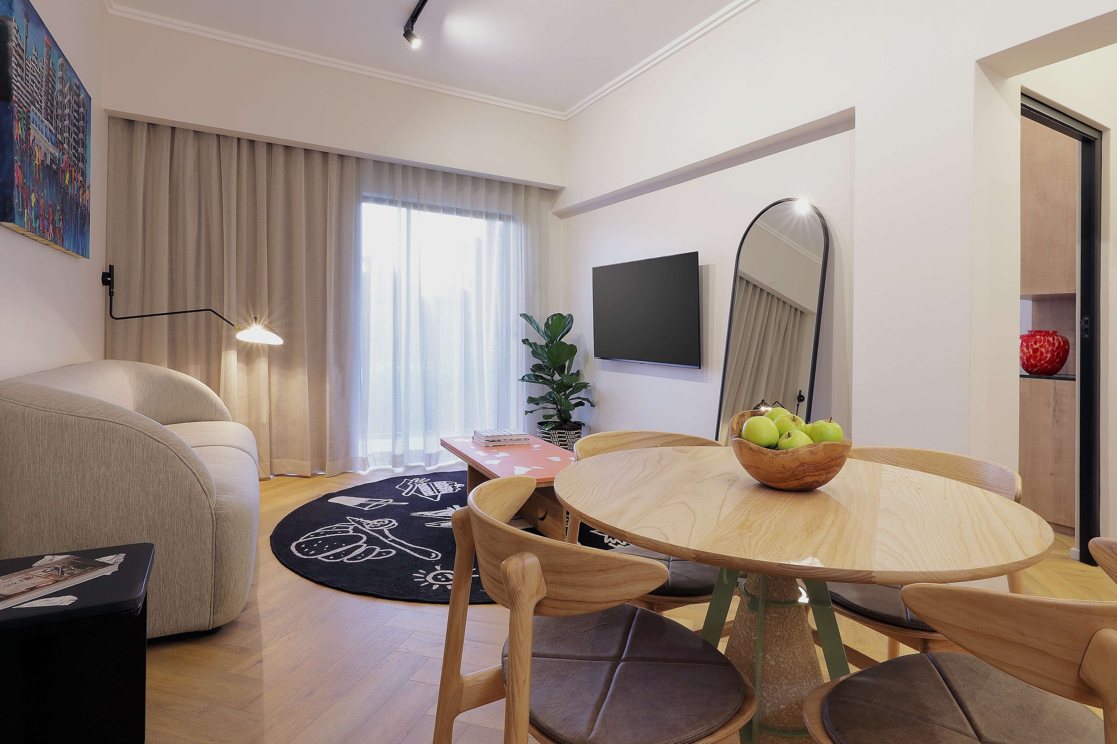Un salon moderne avec un canapé, une table de salle à manger ronde en bois entourée de chaises, un téléviseur à écran plat fixé au mur, une plante en pot et une grande fenêtre avec des rideaux.
