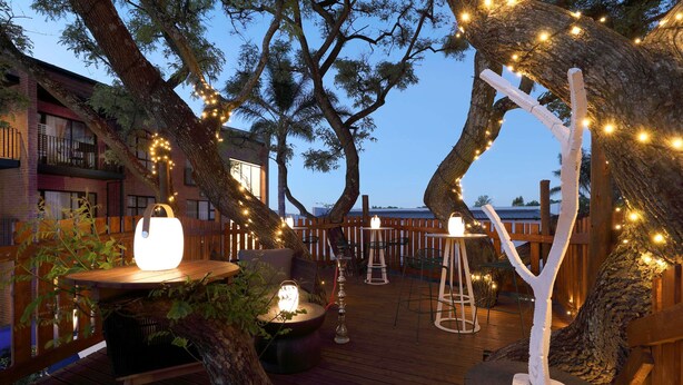 An outdoor deck with wooden flooring, a round table, and chairs, surrounded by trees and string lights. - Hyatt House Johannesburg, Sandton (Sandton)