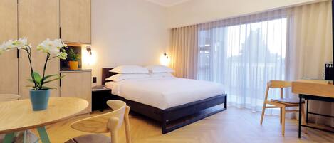 A modern hotel room with a bed, a desk, a chair, a small table, and a potted plant.