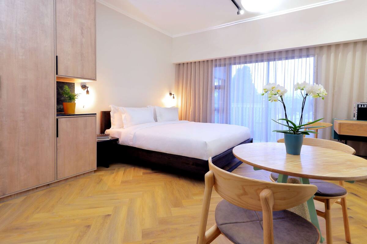 A hotel room with a bed, a desk, a chair, a potted plant, and a window with curtains.