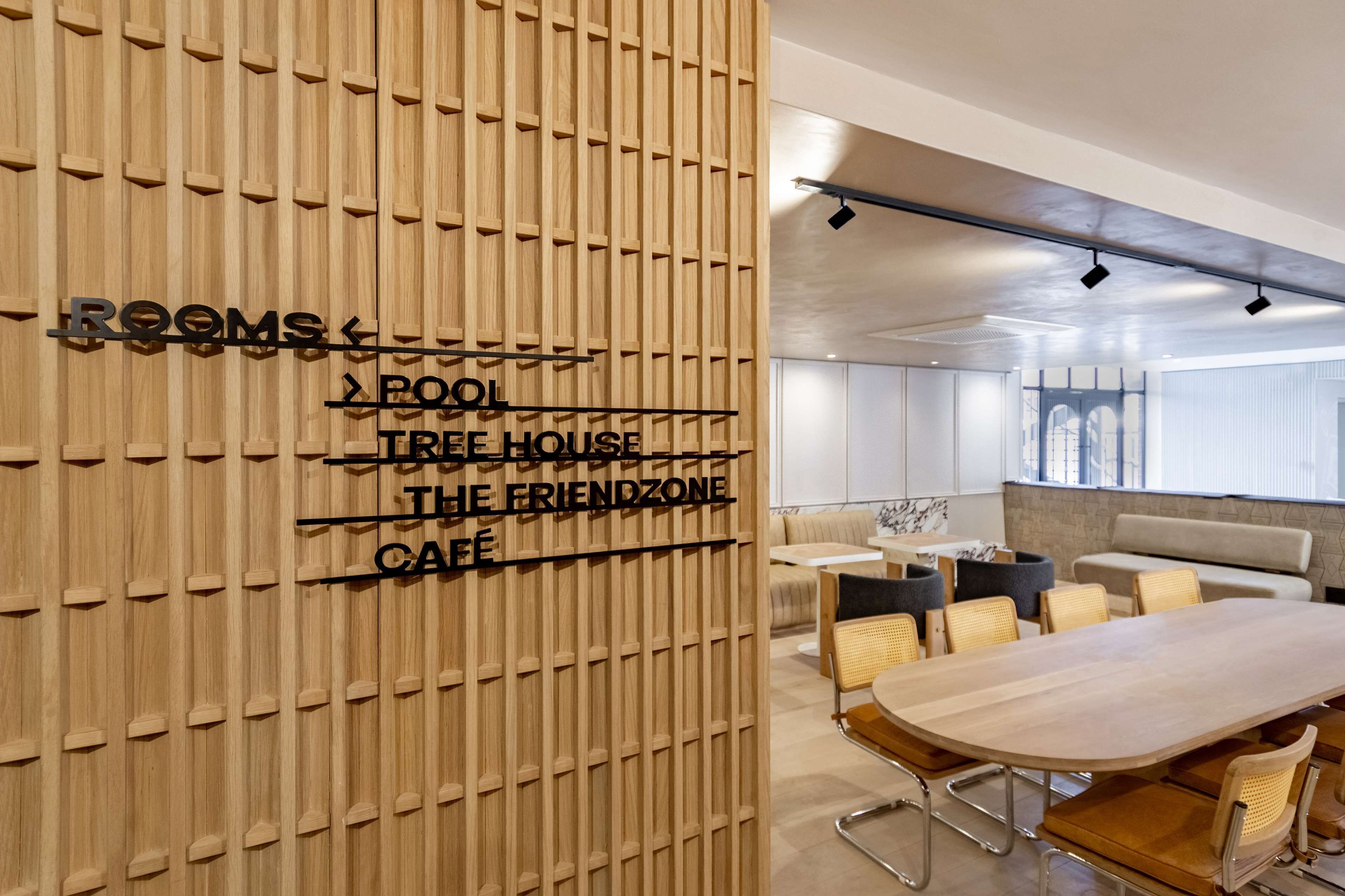 Een modern interieur met een houten scheidingswand waarop zaalnamen staan, een lange tafel met stoelen en een zithoek met banken en kussens.