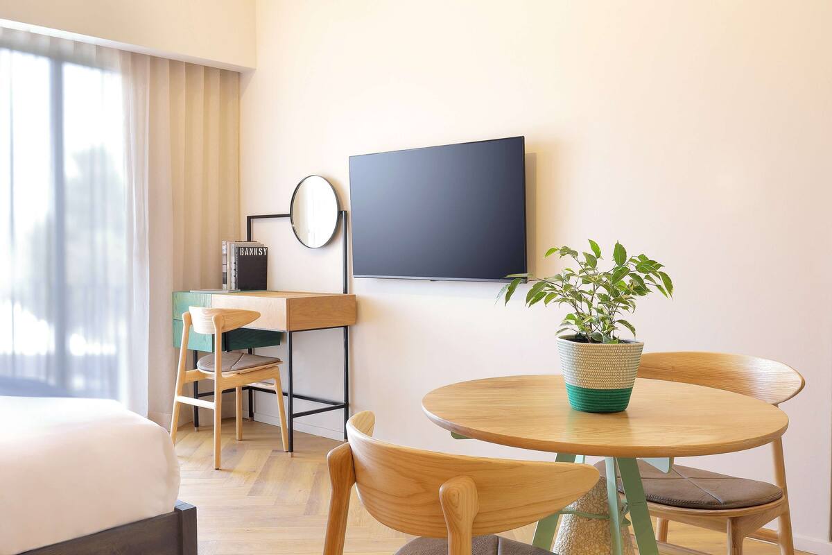 A hotel room with a bed, a desk with a chair, a round table with two chairs, a potted plant, and a flat-screen TV mounted on the wall.