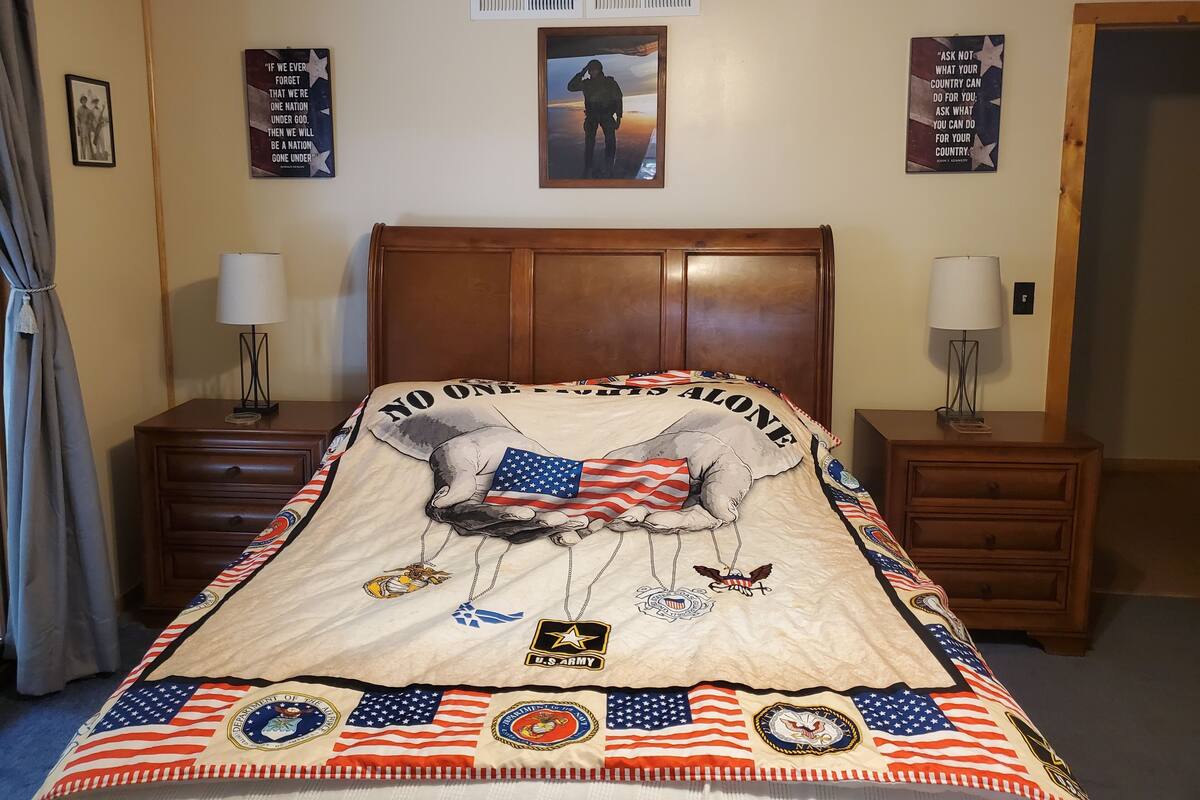 Een slaapkamer met een bed, voorzien van een quilt met militaire en patriottische motieven, twee nachtkastjes met lampen, en motiverende posters aan de muur.