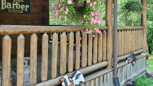 Un porche de madera con una cesta de flores colgante, una cerca de madera y un letrero que dice 'Joy's Barber Shop'.