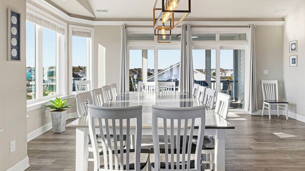 A spacious dining area with a large table and white chairs, a hanging light fixture, and large windows offering a view of the outside. - Great White Shack | 120 ft to the beach | Private Pool, Hot tub (Virginia Beach)