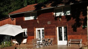 A two-story wooden building with a patio area featuring outdoor furniture and a white umbrella. - A L'orée du Bois - Gîte (Hostun)