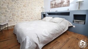 A bedroom with a stone wall, a bed with a patterned bedspread, a nightstand, a lamp, and a wall-mounted shelf. - A L'orée du Bois - Gîte (Hostun)