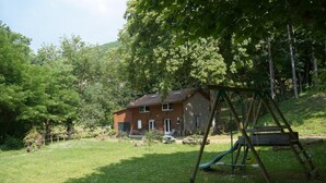 A rustic wooden house with a sloped roof, surrounded by a lush green garden and a swing set. - A L'orée du Bois - Gîte (Hostun)
