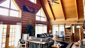 A spacious log cabin interior with a high ceiling, large windows, a ceiling fan, a cozy seating area with a couch and chairs, and a stone fireplace. - Cowboy Cabin in the Mountains - Quiet Getaway (Aguilar)