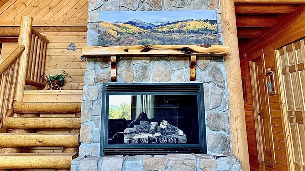 A stone fireplace with a scenic view above it, wooden stairs leading up to it, and a wooden railing on the upper floor. - Cowboy Cabin in the Mountains - Quiet Getaway (Aguilar)