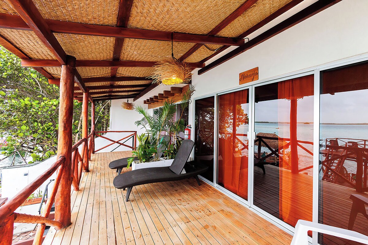 A wooden deck with a thatched roof, a black lounge chair, and a view of the sea through large windows.