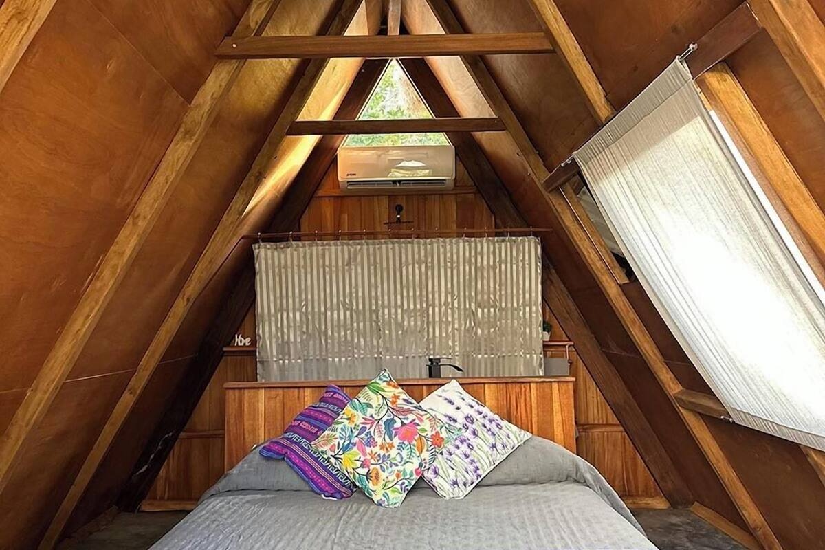 Une chambre mansardée chaleureuse, avec un plafond en pente, un lit recouvert d’une courtepointe violette et une fenêtre agrémentée d’un rideau.