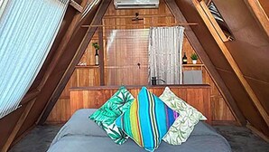 Une chambre aménagée dans les combles, avec un plafond en pente, un lit agrémenté de coussins colorés, une fenêtre avec des rideaux et un petit coin cuisine.
