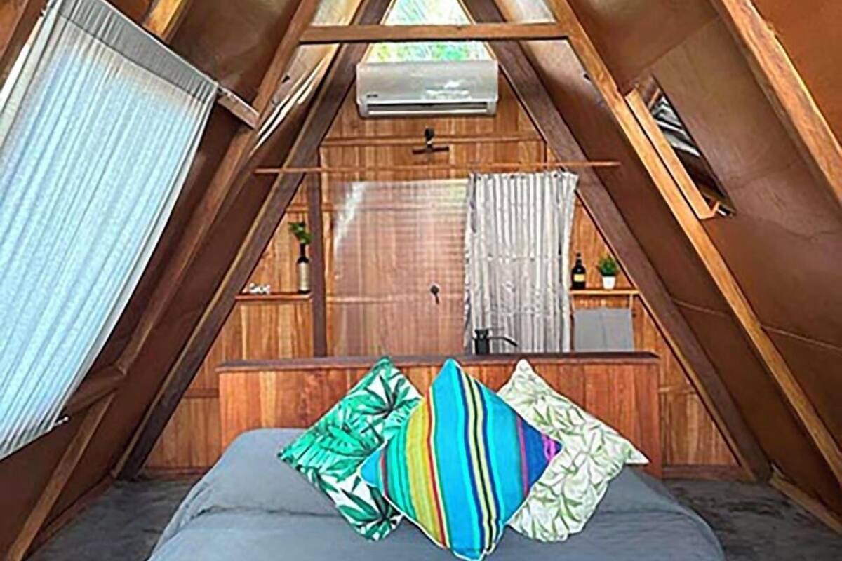 Une chambre aménagée dans les combles, avec un plafond en pente, un lit agrémenté de coussins colorés, une fenêtre avec des rideaux et un petit coin cuisine.