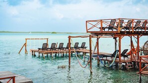 Une jetée en bois avec des chaises longues qui s’étendent au-dessus d’une étendue d’eau calme, un petit bateau au loin et un ciel dégagé.