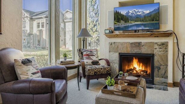 A living room with a fireplace, a television mounted above it, and a large window overlooking a building. - Trade the Office for Mountains at Foxpoint Townhouse 6180 (Park City)