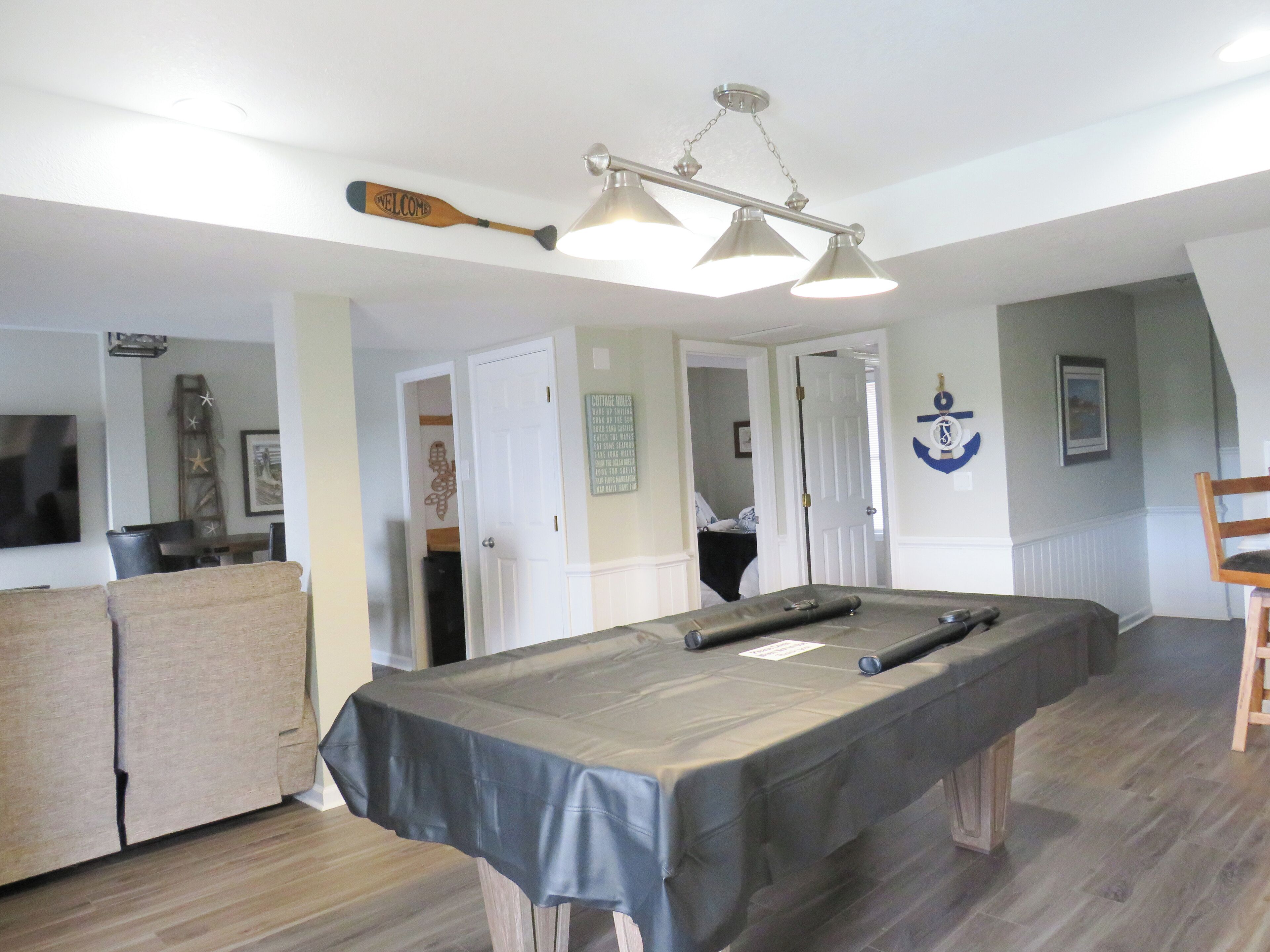 Une table de billard recouverte d’une nappe, un queue de billard et un luminaire suspendu dans une pièce au plancher en bois et aux murs blancs.