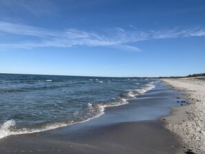 Ein Strand mit sanft anschwellenden Wellen, einem klaren Himmel und einer fernen Uferlinie mit Vegetation.