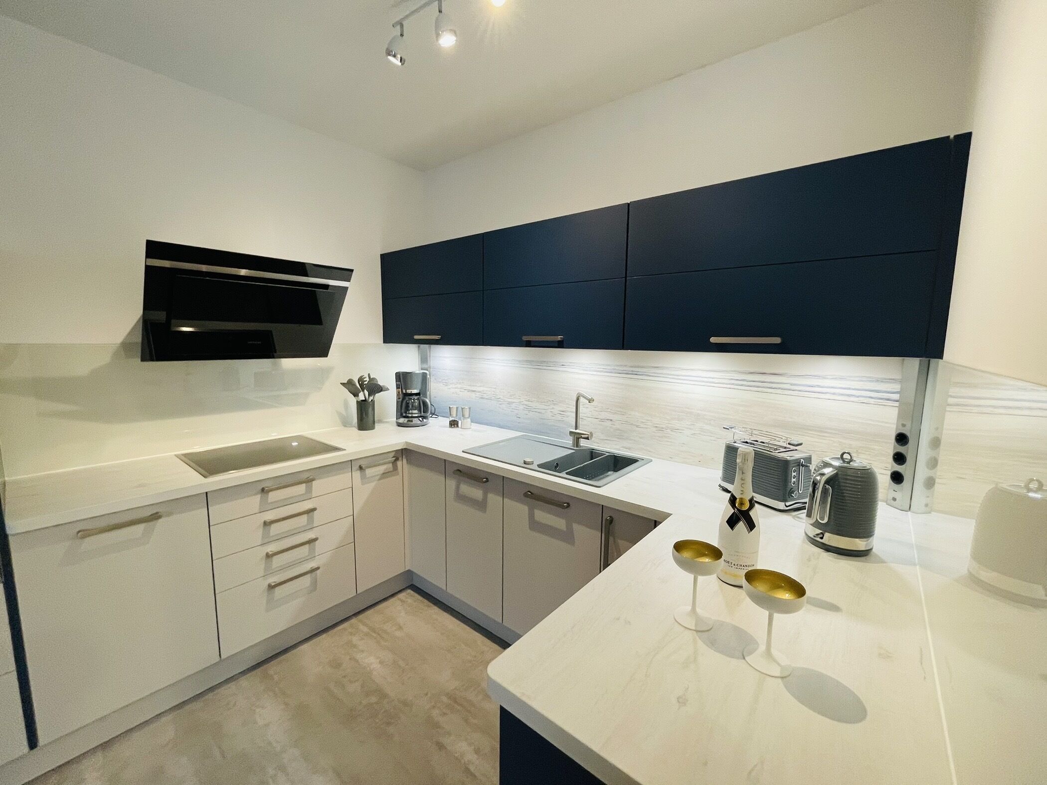 Una cocina moderna con encimeras blancas, un microondas integrado, una cafetera y un juego de copas de vino sobre la encimera.