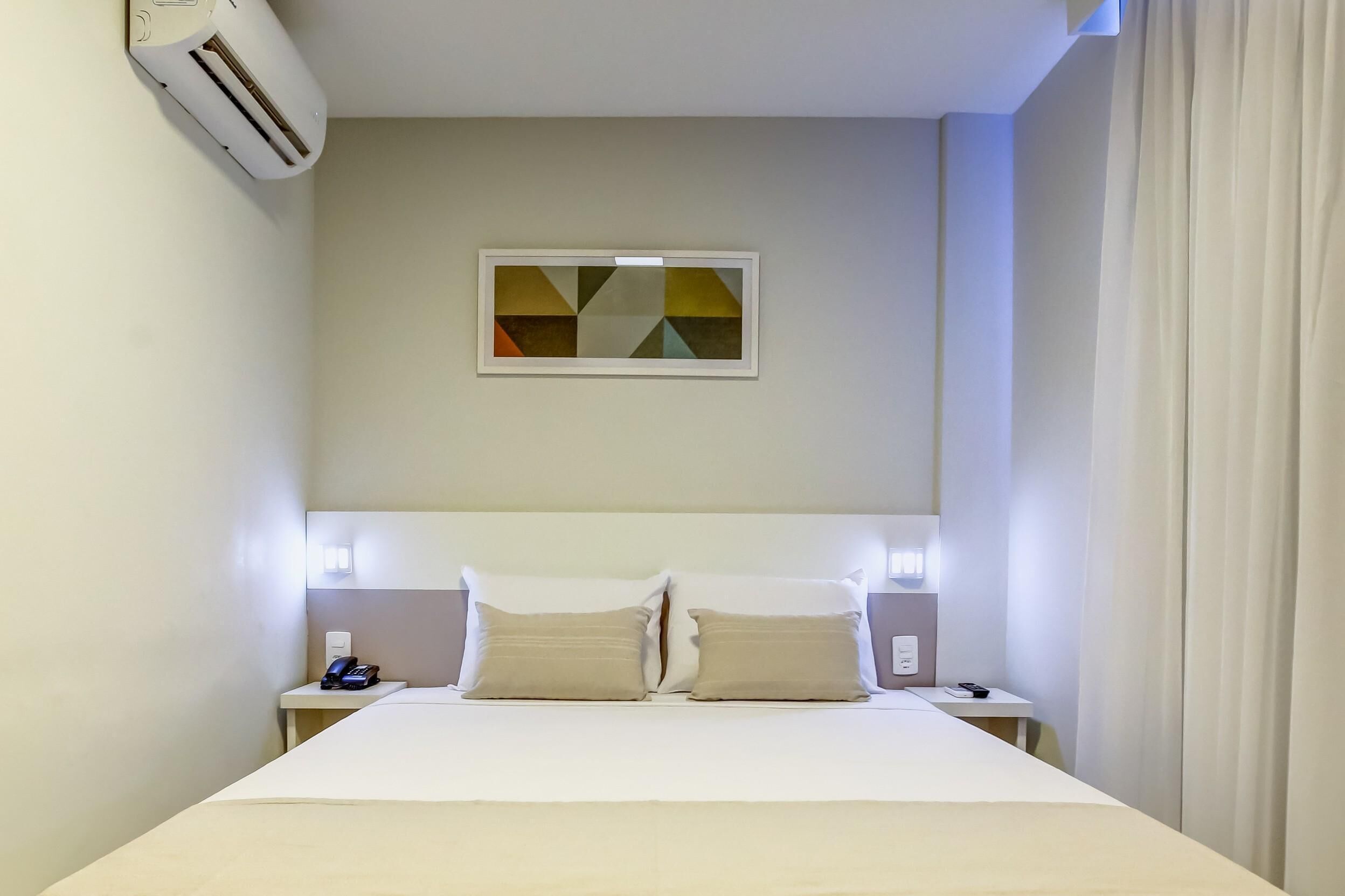 Une chambre d’hôtel avec un lit, deux tables de chevet, un climatiseur fixé au mur, une œuvre d’art géométrique accrochée au mur et un rideau à la fenêtre.