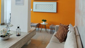 Una sala moderna con un sofá blanco, una mesa de centro con objetos decorativos y dos obras de arte enmarcadas sobre una pared naranja.