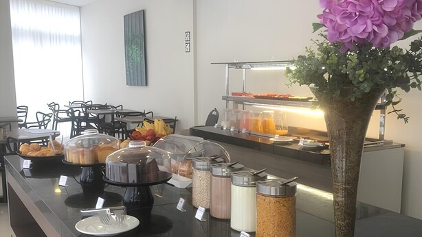 Zona de desayuno tipo buffet con diferentes opciones de alimentos, un jarrón con flores y un área de estar con mesas y sillas.