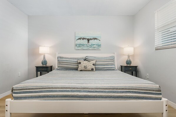 A bed with a striped bedspread, two bedside tables with lamps, a wall-mounted picture of a whale, and a window with blinds.