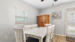 A dining room with a white table and chairs, a wooden cabinet, a ceiling fan, and a painting on the wall. - 2-Bedroom Condo with Waterview Balcony - Tarpon - Welcome to our stunning two-bedroom condo room with a balcony! (Matlacha)