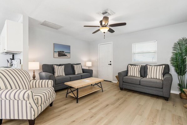 A living room with two grey sofas, a striped armchair, a wooden coffee table, a ceiling fan, and a large potted plant. - 2-Bedroom Condo with Waterview Balcony - Tarpon - Welcome to our stunning two-bedroom condo room with a balcony! (Matlacha)