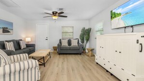 A living room with a grey sofa, a striped armchair, a wooden coffee table, a ceiling fan, a TV on the wall, and a white cabinet. - 2-Bedroom Condo with Waterview Balcony - Tarpon - Welcome to our stunning two-bedroom condo room with a balcony! (Matlacha)