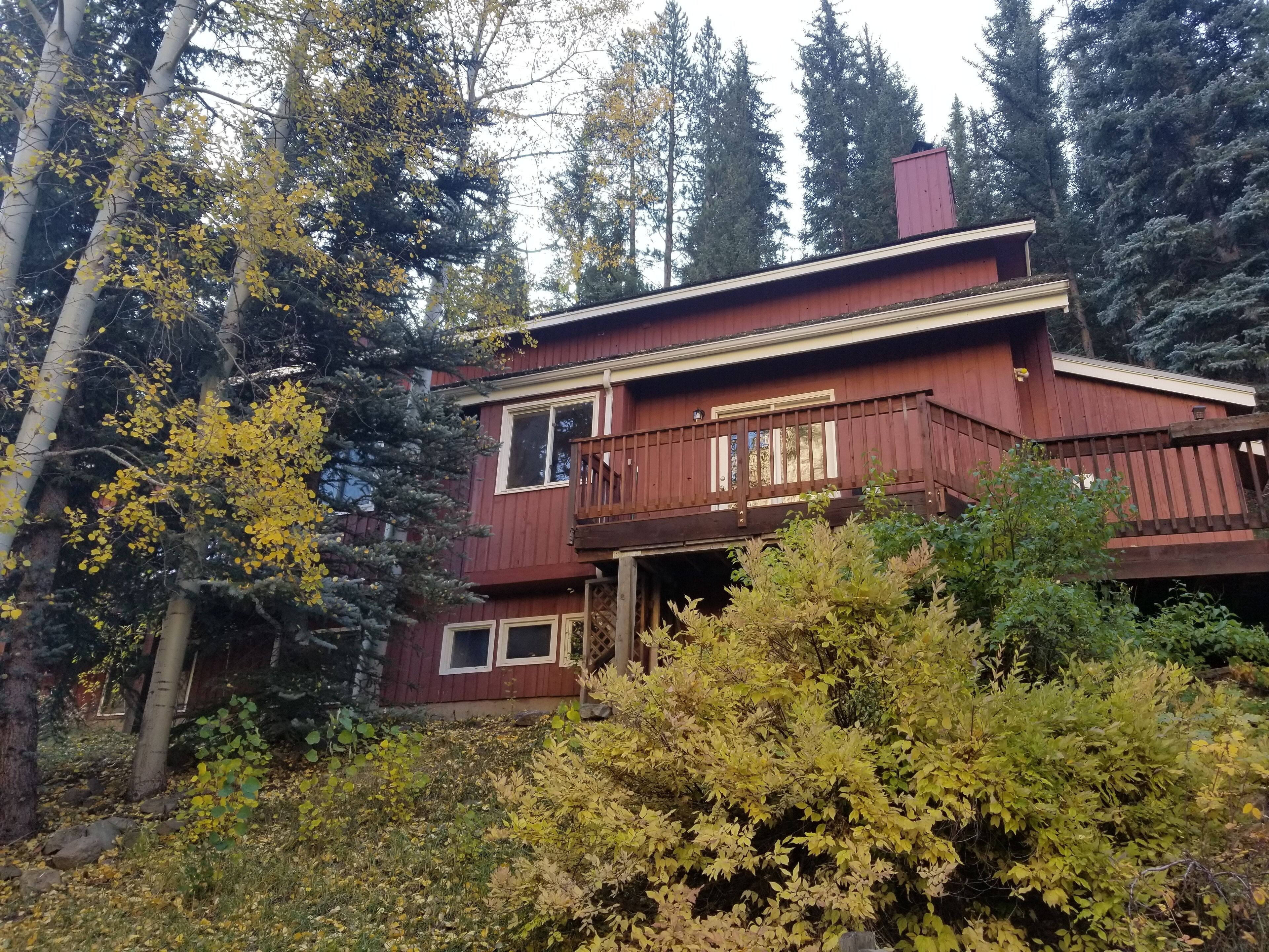 A two-story wooden cabin with a balcony, surrounded by dense forest and autumn foliage.