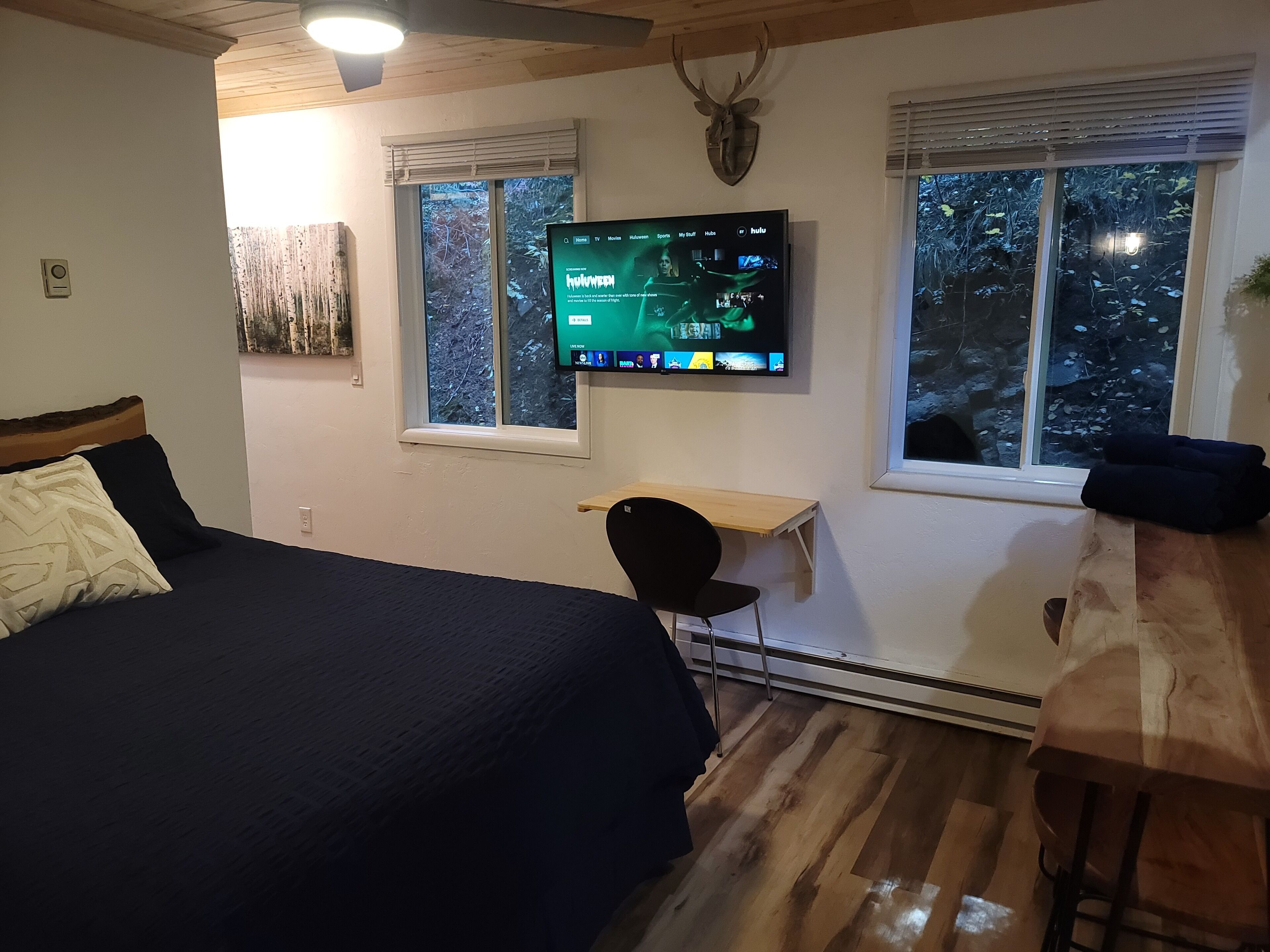 A bedroom with a bed, a desk, a chair, a mounted TV, and windows with a view of trees.