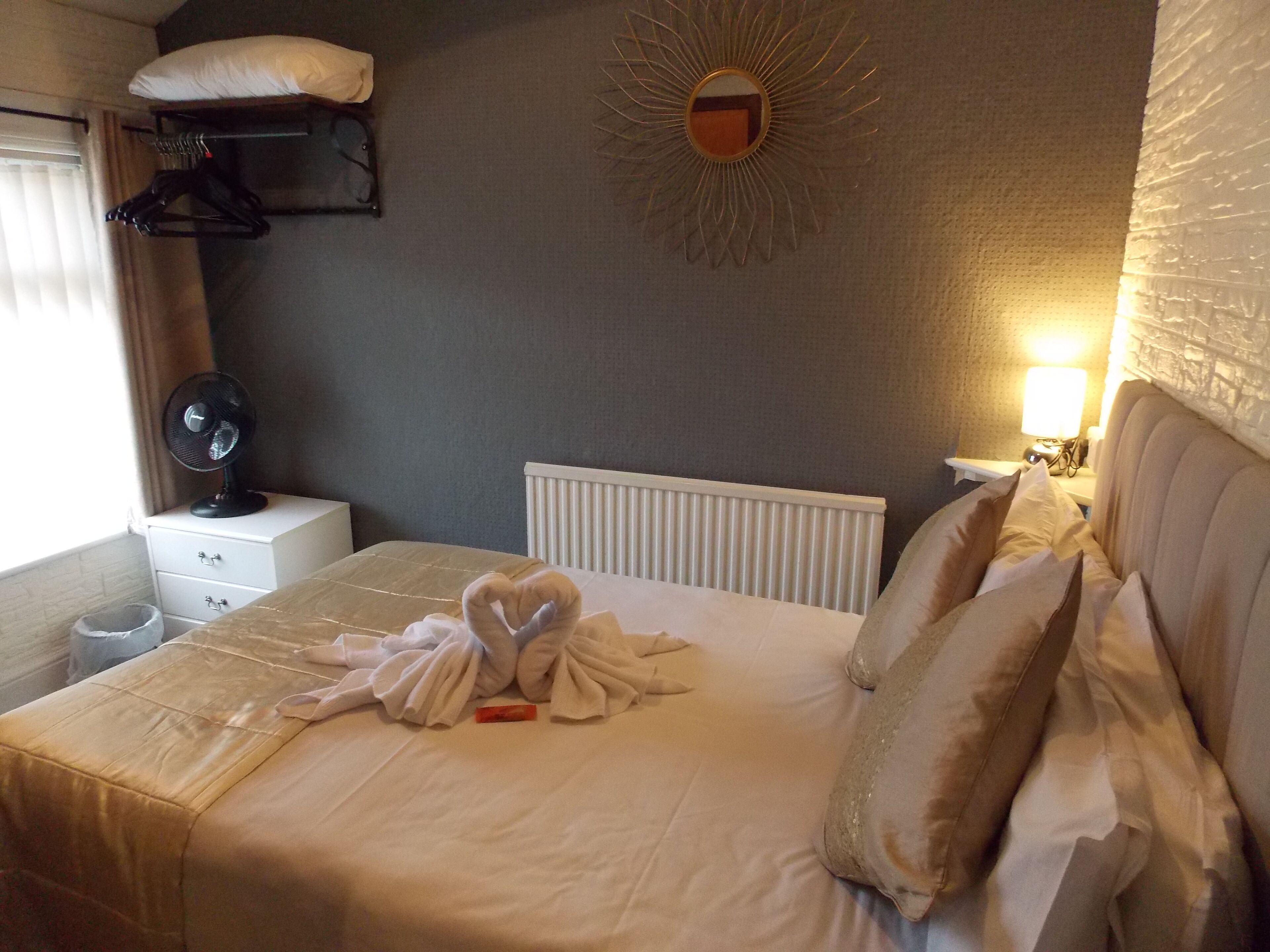 Una cama bien hecha con una decoración de toallas con forma de corazón, una mesita de noche con un ventilador, un espejo en la pared y un radiador.