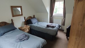 A bedroom with two single beds, a nightstand with a lamp, a wooden headboard, a window with curtains, and a radiator. - The Cobbler Cottage - Loch Lomond and Arrochar Alps (Arrochar)