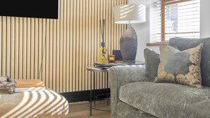 A modern living room with a grey sofa, a patterned ottoman, a lamp, and a small table with a book and a bottle. - Coach House (Bridgend)