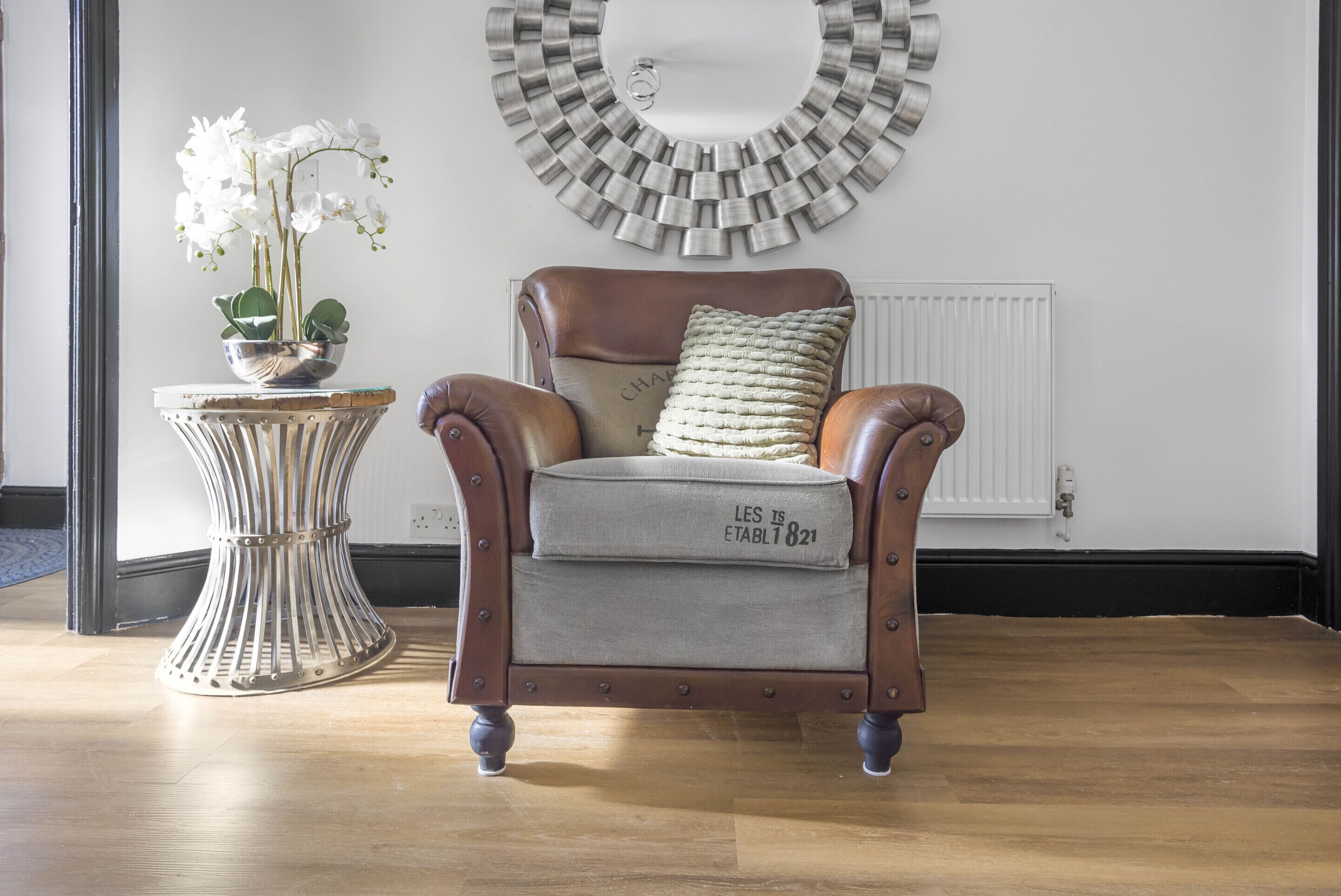 A brown leather armchair with a cushion labeled "LES ESTABLIS 821" and a woven pillow, positioned in front of a white radiator and a wall-mounted mirror.