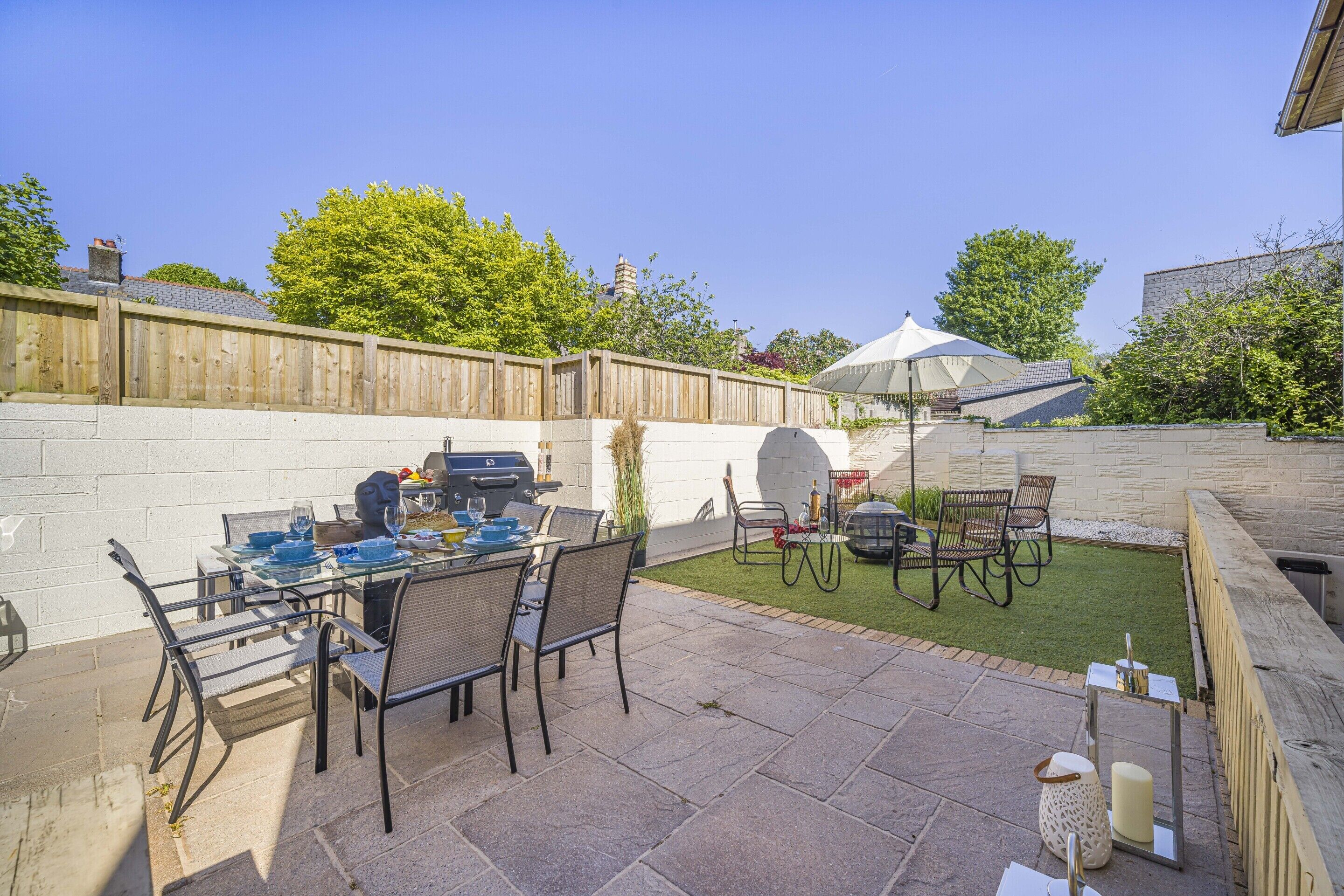 An outdoor patio with a dining table set for four, surrounded by chairs. There is a barbecue grill and a covered seating area.